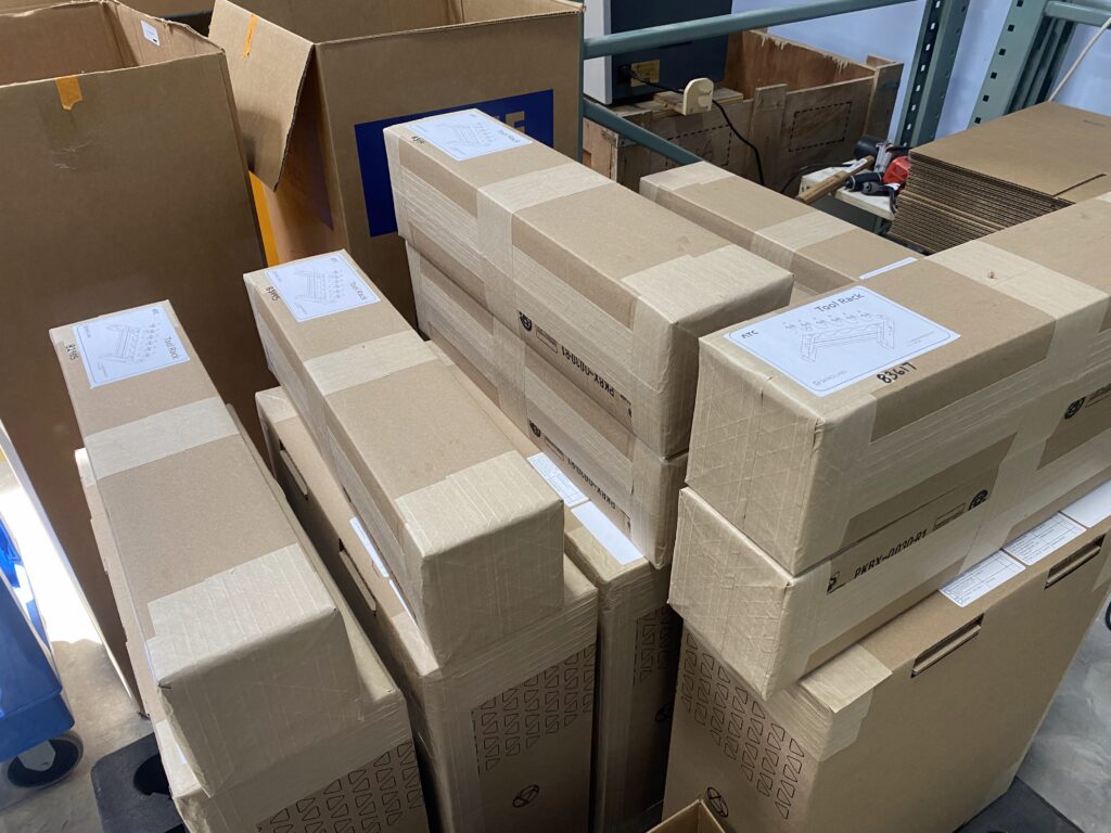 Piles of brown cardboard boxes stacked in a warehouse storage area, some with labels and printed diagrams on the flaps.