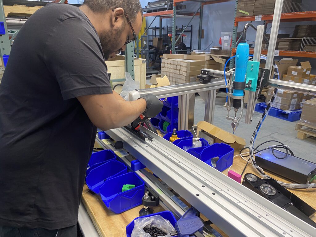 Worker in a black shirt wearing gloves, operating a handheld tool along a metal assembly line in a warehouse; blue storage bins and cardboard boxes visible in the background.