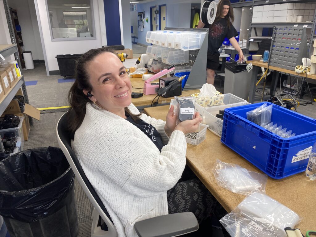 Smiling woman seated at a cluttered workshop table, holding clear plastic parts organizers with small bins nearby.
