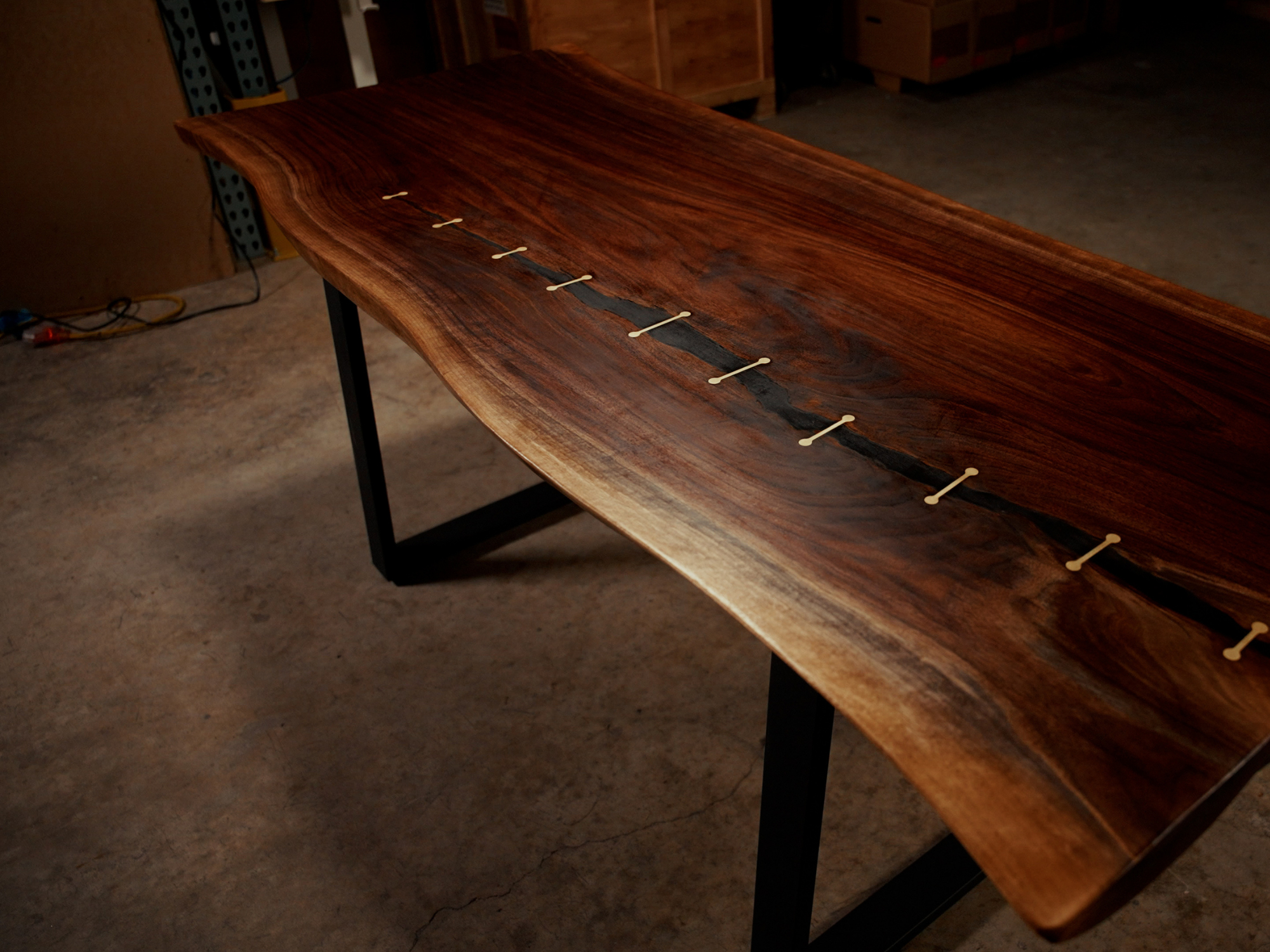 Live-edge wooden table with a dark resin river and small white inlays along the seam in a workshop setting.