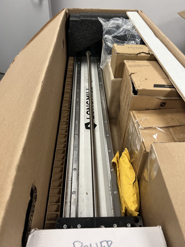 Industrial machinery parts packed inside a large cardboard crate: a silver linear rail with bolts and a coiled rod, surrounded by foam, boxes, and packing material