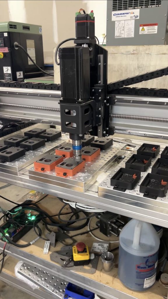 Automated CNC/assembly station with a vertical robotic spindle above orange clamping blocks on a perforated work table of metal parts and cables nearby.