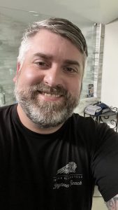 IMG_2970 | Sienci Labs Close-up of a man with gray beard smiling, taking a selfie in a bathroom; wearing a black T-shirt with Daytona Beach graphic.